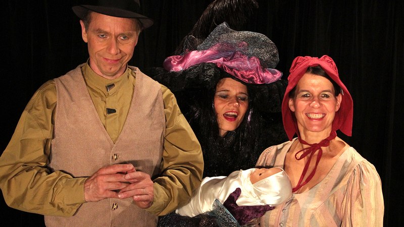 Drei Personen in Kostümen auf einer Bühne. Eine Frau hält ein Baby, eine andere trägt einen großen Hut. Der Mann trägt einen Hut und eine Weste., © Galli Theater Backnang Drei Personen in Kostümen auf einer Bühne. Eine Frau hält ein Baby, eine andere trägt einen großen Hut. Der Mann trägt einen Hut und eine Weste., © Galli Theater Backnang