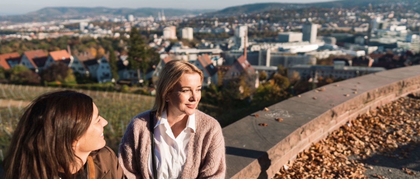 Zwei Frauen sitzen auf einer Mauer, genießen die Aussicht auf eine Stadt mit Hügeln im Hintergrund. Herbstlaub liegt auf der Mauer., © SMG, wpsteinheisser Zwei Frauen sitzen auf einer Mauer, genießen die Aussicht auf eine Stadt mit Hügeln im Hintergrund. Herbstlaub liegt auf der Mauer., © SMG, wpsteinheisser
