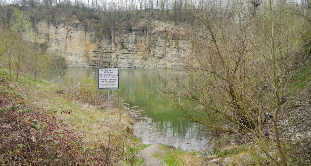 Ein kleiner See mit steilen Felswänden, umgeben von Bäumen. Ein Warnschild weist auf Gefahren hin. Der Himmel ist bewölkt., © Hans Sukowski Ein kleiner See mit steilen Felswänden, umgeben von Bäumen. Ein Warnschild weist auf Gefahren hin. Der Himmel ist bewölkt., © Hans Sukowski