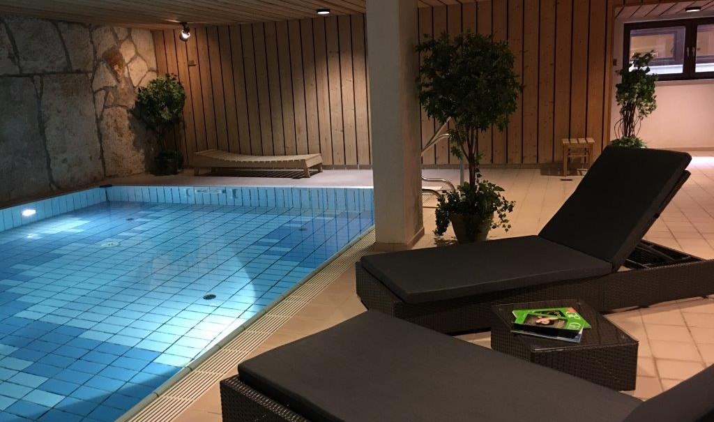 Ein beheizter Indoor-Pool mit blauen Fliesen, umgeben von Liegestühlen und Pflanzen. Die Wände sind aus Holz und Stein., © TOMAS Ein beheizter Indoor-Pool mit blauen Fliesen, umgeben von Liegestühlen und Pflanzen. Die Wände sind aus Holz und Stein., © TOMAS