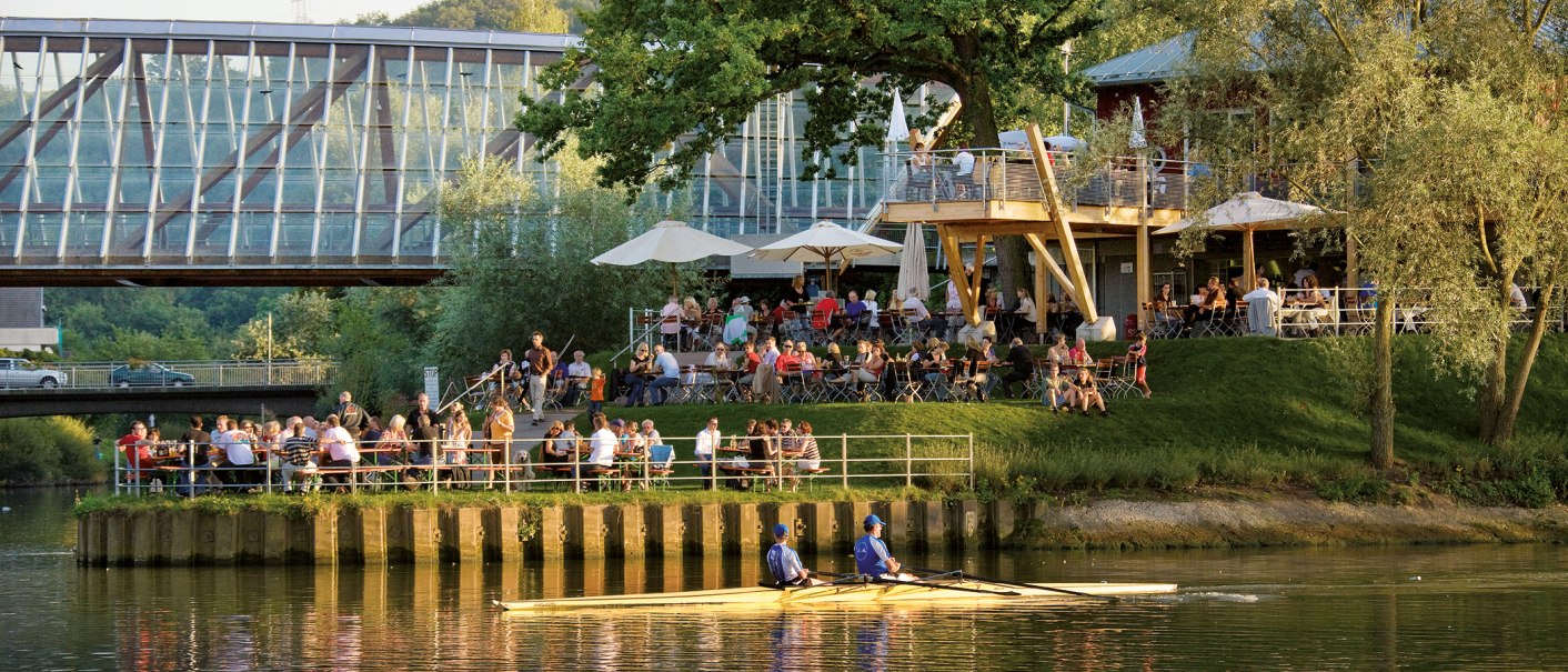 Menschen genießen das Wetter in einem Café am Flussufer, während ein Ruderboot vorbeifährt. Im Hintergrund ist eine moderne Brücke zu sehen., © Michael Fuchs