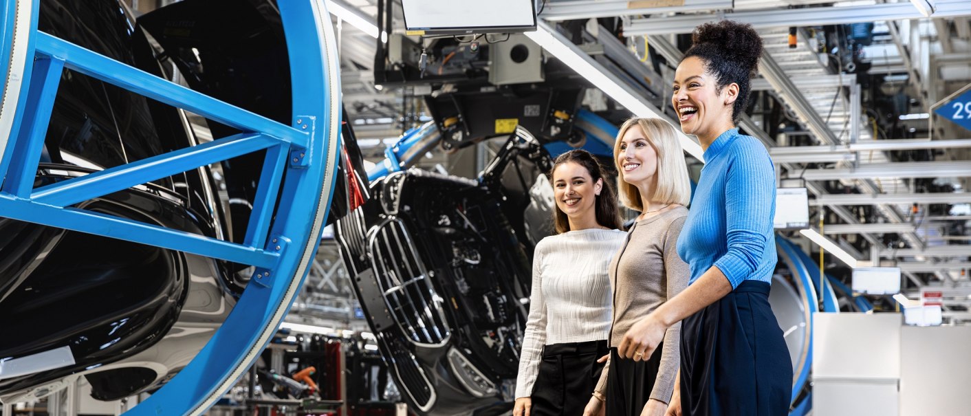 Drei Frauen stehen lächelnd in einer modernen Fabrikhalle. Im Hintergrund sind Autoteile und industrielle Anlagen zu sehen., © Mercedes-Benz AG Drei Frauen stehen lächelnd in einer modernen Fabrikhalle. Im Hintergrund sind Autoteile und industrielle Anlagen zu sehen., © Mercedes-Benz AG
