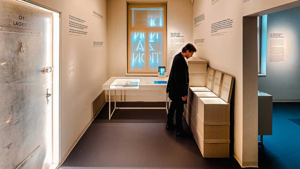 Ein Museumsraum mit einer Person, die Exponate betrachtet. An der Wand sind Texte und ein beleuchtetes Fenster zu sehen. Links eine Tür mit der Aufschrift '01 Lager'., © Haus der Geschichte Baden-Württemberg