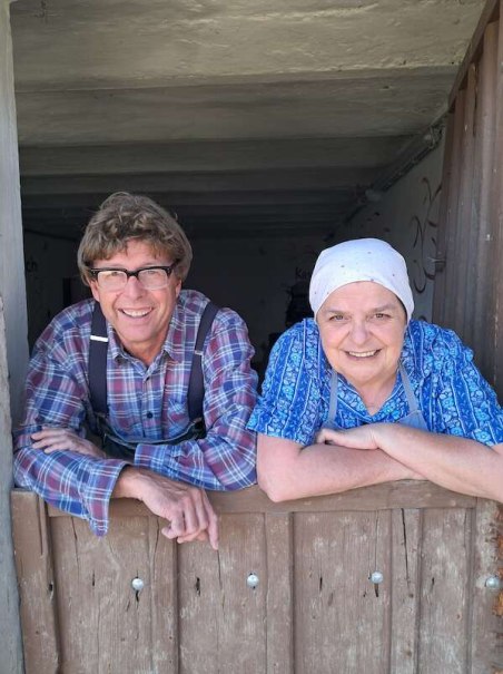 Zwei Personen lehnen sich lächelnd über eine Holztür. Eine Person trägt ein kariertes Hemd, die andere ein blaues Kopftuch und eine Schürze., © SBEntertainment GmbH & Co. KG Zwei Personen lehnen sich lächelnd über eine Holztür. Eine Person trägt ein kariertes Hemd, die andere ein blaues Kopftuch und eine Schürze., © SBEntertainment GmbH & Co. KG