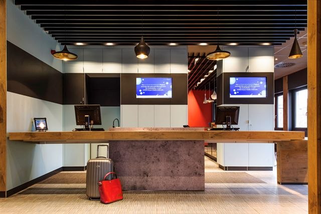 Moderne Hotelrezeption mit zwei Bildschirmen, Gepäckstücken und stilvoller Beleuchtung., © TOMAS