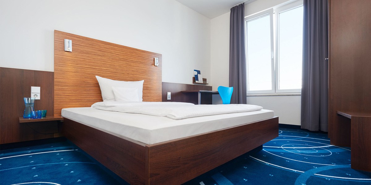 Modernes Hotelzimmer mit Einzelbett, Holzmöbeln, blauem Teppich und großem Fenster mit grauen Vorhängen., © TOMAS