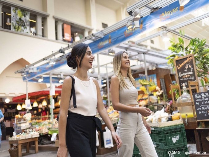 Zwei Frauen schlendern lachend durch die Stuttgarter Markthalle, umgeben von bunten Marktständen und frischen Lebensmitteln., © Stuttgart Marketing GmbH Zwei Frauen schlendern lachend durch die Stuttgarter Markthalle, umgeben von bunten Marktständen und frischen Lebensmitteln., © Stuttgart Marketing GmbH