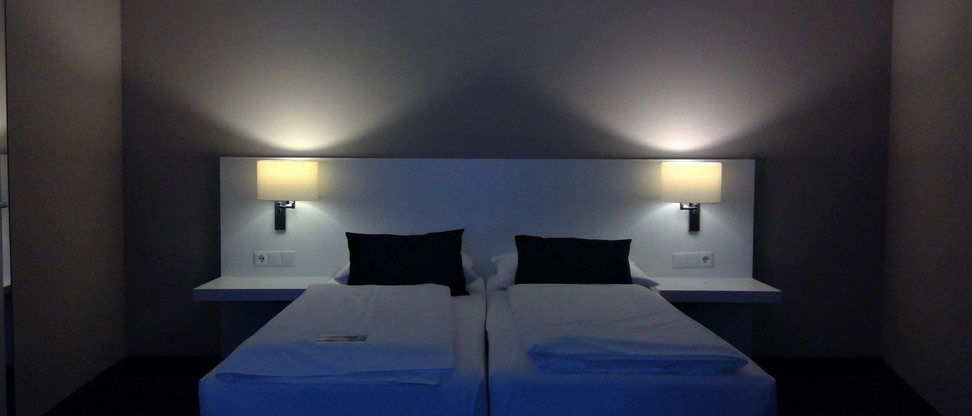 Zwei Einzelbetten mit weißen Laken und schwarzen Kissen in einem Hotelzimmer. An der Wand sind zwei Lampen angebracht, die warmes Licht spenden., © relexa Waldhotel Schatten Zwei Einzelbetten mit weißen Laken und schwarzen Kissen in einem Hotelzimmer. An der Wand sind zwei Lampen angebracht, die warmes Licht spenden., © relexa Waldhotel Schatten
