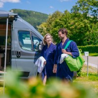 Ein Paar in blauen Bademänteln steht lächelnd vor einem modernen Wohnmobil auf einem sonnigen Campingplatz., © Thomas Niedermüller Ein Paar in blauen Bademänteln steht lächelnd vor einem modernen Wohnmobil auf einem sonnigen Campingplatz., © Thomas Niedermüller