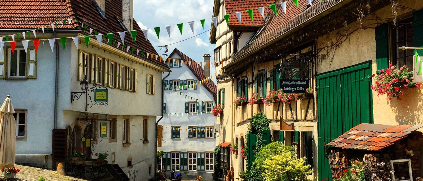 Malerische Altstadtgasse mit Kopfsteinpflaster, Fachwerkhäusern und bunten Wimpeln. Blumen schmücken die Fassaden, sonniges Wetter., © Nürtingen Malerische Altstadtgasse mit Kopfsteinpflaster, Fachwerkhäusern und bunten Wimpeln. Blumen schmücken die Fassaden, sonniges Wetter., © Nürtingen