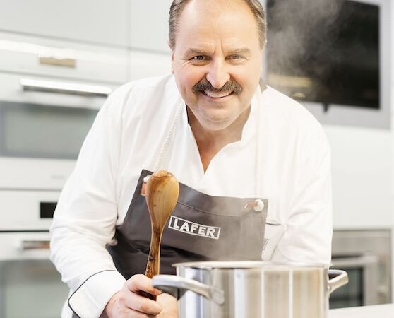 Ein Koch in einer modernen K&uuml;che r&uuml;hrt mit einem Holzl&ouml;ffel in einem Topf. Er tr&auml;gt eine Sch&uuml;rze mit der Aufschrift 'Lafer'., &copy; Stadthalle Leonberg