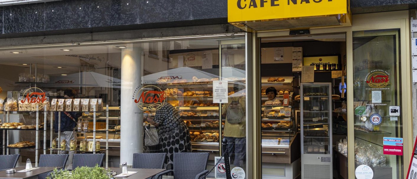 Außenansicht des Cafe Nast mit großen Fenstern, durch die Backwaren und Kunden zu sehen sind. Ein gelbes Schild mit dem Café-Namen hängt über dem Eingang., © SMG, Sarah Schmid Außenansicht des Cafe Nast mit großen Fenstern, durch die Backwaren und Kunden zu sehen sind. Ein gelbes Schild mit dem Café-Namen hängt über dem Eingang., © SMG, Sarah Schmid