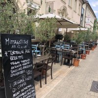 Ein Straßencafé mit Tischen und Stühlen unter Sonnenschirmen. Eine Menütafel bietet Tortellini und Fleischgerichte an. Olivenbäume in Töpfen säumen den Weg., © Antipasto Wein und mehr