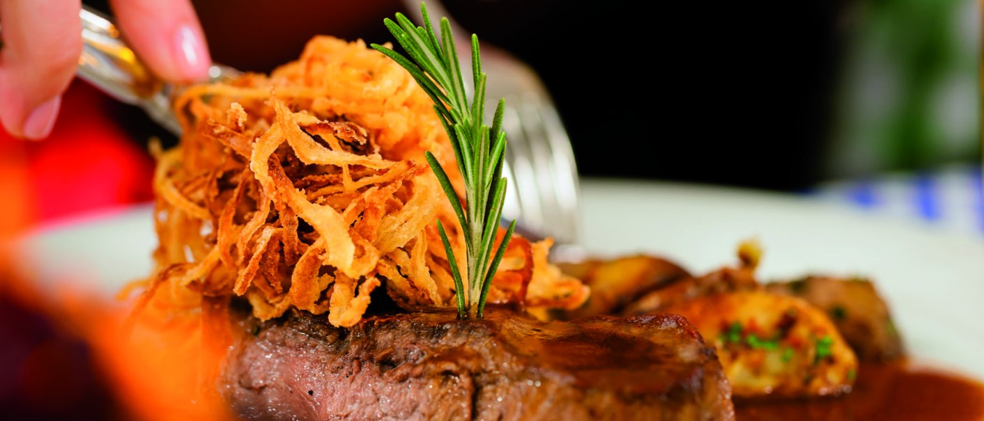 Zwiebelrostbraten mit knusprigen Zwiebeln und Rosmarin garniert. Im Hintergrund sind Beilagen zu sehen., © Kzenon-Fotolia