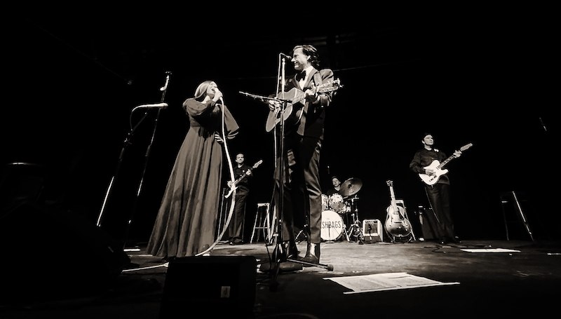 Schwarz-weiß Bild einer Band auf der Bühne. Sängerin und Sänger mit Gitarre im Vordergrund, Schlagzeuger und Gitarristen im Hintergrund., © Good Concert GmbH