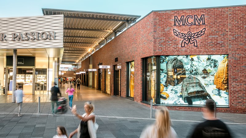 Einkaufsstraße in der Outletcity Metzingen mit Geschäften wie MCM. Menschen schlendern vorbei, einige sind in Bewegung unscharf., © OUTLETCITY AG Einkaufsstraße in der Outletcity Metzingen mit Geschäften wie MCM. Menschen schlendern vorbei, einige sind in Bewegung unscharf., © OUTLETCITY AG