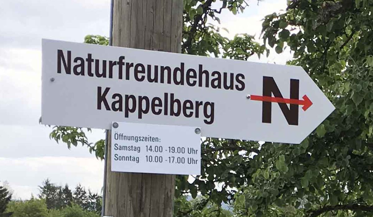 Wegweiser zum Naturfreundehaus Kappelberg mit Öffnungszeiten für Samstag und Sonntag, montiert an einem Holzpfosten vor Bäumen. Wegweiser zum Naturfreundehaus Kappelberg mit Öffnungszeiten für Samstag und Sonntag, montiert an einem Holzpfosten vor Bäumen.