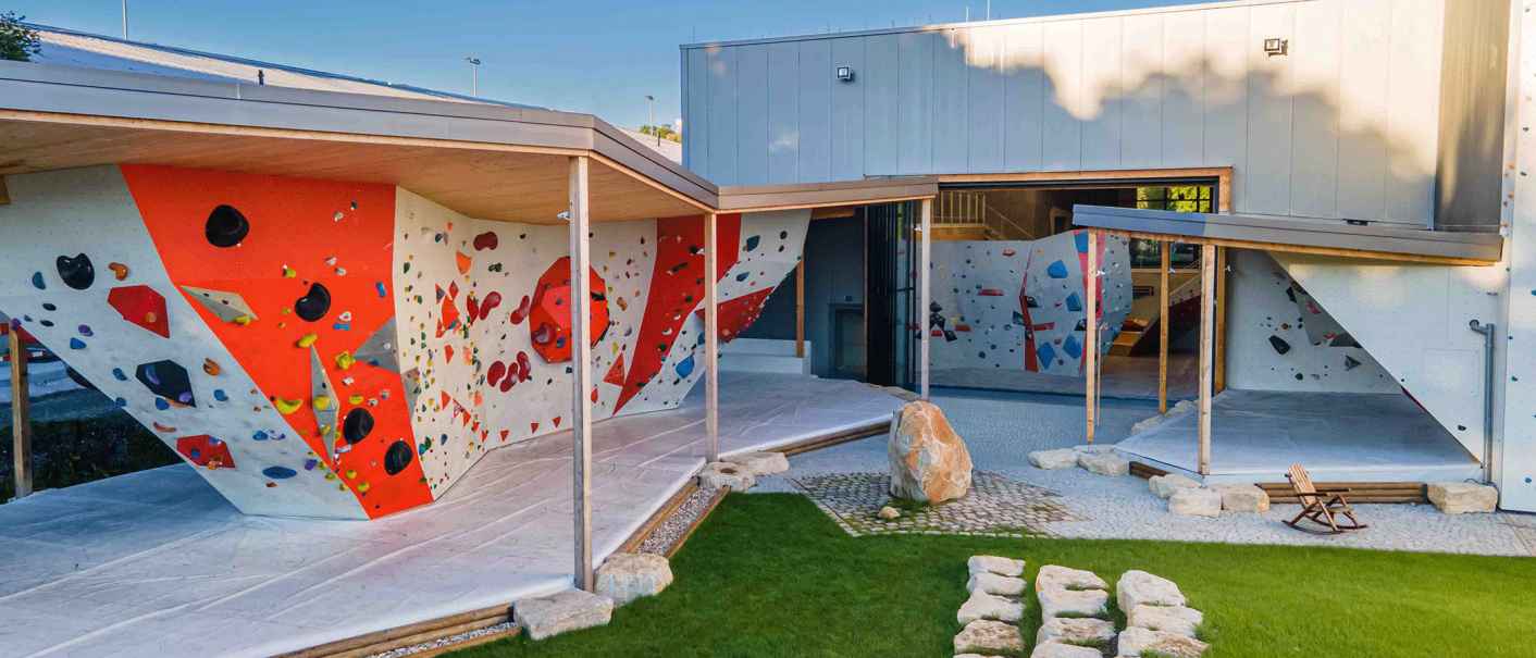 Moderne Kletterhalle mit bunten Kletterwänden im Freien, umgeben von einem gepflegten Garten mit Steinen und Rasen., © Roccadion, Böblingen Moderne Kletterhalle mit bunten Kletterwänden im Freien, umgeben von einem gepflegten Garten mit Steinen und Rasen., © Roccadion, Böblingen