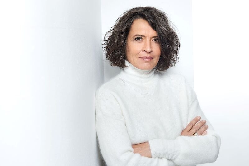 Eine Frau mit lockigem Haar in einem weißen Rollkragenpullover lehnt entspannt an einer weißen Wand und schaut in die Kamera., © Kulturbüro Schwäbisch Gmünd