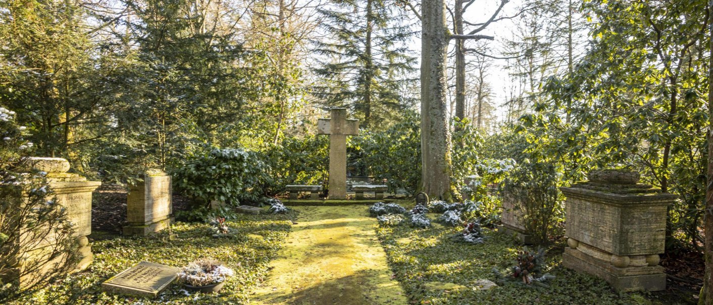 Ein ruhiger Friedhof mit einem großen Kreuz in der Mitte, umgeben von Bäumen und grüner Vegetation. Sonnenlicht fällt durch die Blätter., © Stuttgart-Marketing GmbH, Sarah Schmid
