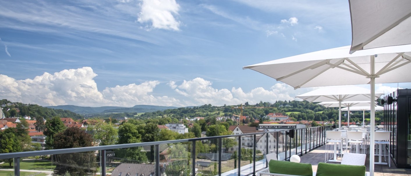 Rooftop-Bar mit weißen Sonnenschirmen und grünen Kissen, Blick auf eine Stadtlandschaft mit Hügeln im Hintergrund., © Hotel am Remspark Rooftop-Bar mit weißen Sonnenschirmen und grünen Kissen, Blick auf eine Stadtlandschaft mit Hügeln im Hintergrund., © Hotel am Remspark