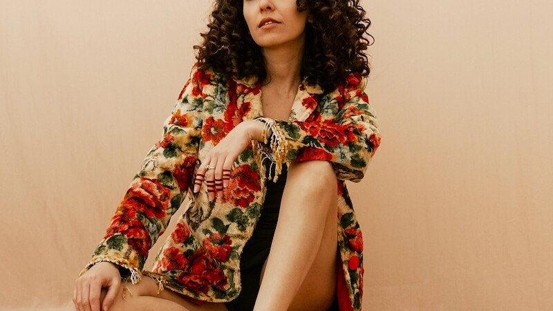 Eine Frau mit lockigem Haar sitzt in einem geblümten Mantel auf einem beigen Hintergrund. Sie wirkt entspannt und selbstbewusst., © Kulturhaus Schwanen