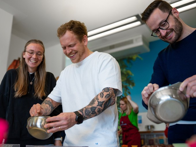 Drei Personen lachen und gie&szlig;en Schokolade in Formen. Im Hintergrund arbeitet eine weitere Person. Alle wirken fr&ouml;hlich und konzentriert., &copy; Alfred Ritter GmbH &amp; Co. KG | Waldenbuch