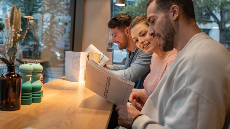 Drei Personen sitzen an einem Tisch in einem Restaurant und lesen die Speisekarte. Im Hintergrund ist ein Fenster mit Blick nach draußen., © Stuttgart-Marketing GmbH, Martina Denker