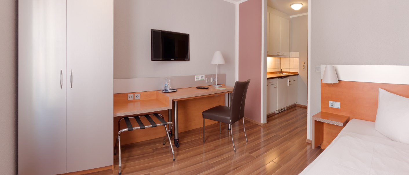 Modernes Einzelzimmer mit Bett, Schreibtisch, Stuhl, Fernseher und Kleiderschrank. Helle Holzmöbel und Laminatboden, kleine Küchenzeile im Hintergrund., © attimo Hotel Stuttgart
