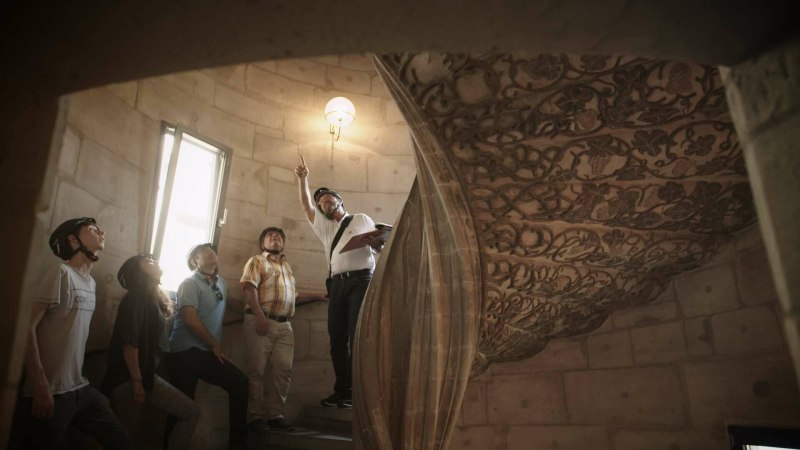 Eine Gruppe von Menschen mit Helmen steht in einem historischen Geb&auml;ude. Ein F&uuml;hrer zeigt auf die kunstvoll verzierte Decke.