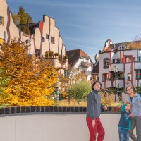 Menschen stehen vor dem farbenfrohen Hundertwasser-Wohnhaus in Plochingen. Die Fassade ist wellenförmig und bunt gestaltet, umgeben von Herbstbäumen., © Bildergalerie Attilla Menschen stehen vor dem farbenfrohen Hundertwasser-Wohnhaus in Plochingen. Die Fassade ist wellenförmig und bunt gestaltet, umgeben von Herbstbäumen., © Bildergalerie Attilla