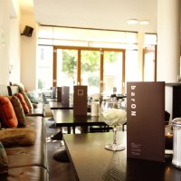 Gemütliches Café mit Kissen auf Bänken, Speisekarten auf Tischen und einem Glas mit Blüten. Große Fenster lassen viel Licht herein., © Andreas Ebert - BARON