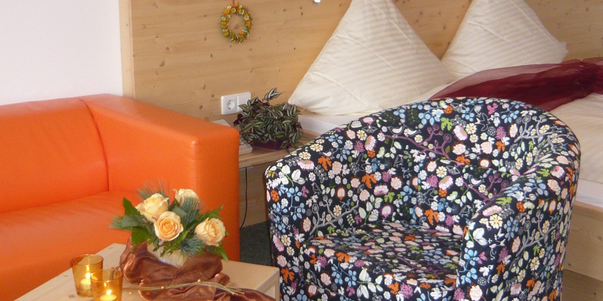Gemütliches Doppelzimmer mit oranger Couch, floralem Sessel, Blumenstrauß und Bett im Hintergrund., © Hotel Hochwald