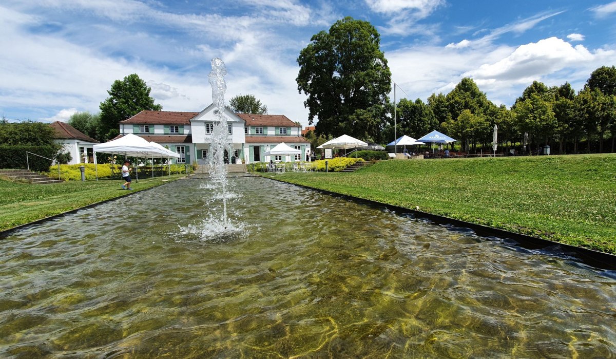Weißes Gebäude mit rotem Dach hinter Wasserbecken und Springbrunnen, umgeben von grüner Wiese und Bäumen unter blauem Himmel., © Kulturamt Plochingen Weißes Gebäude mit rotem Dach hinter Wasserbecken und Springbrunnen, umgeben von grüner Wiese und Bäumen unter blauem Himmel., © Kulturamt Plochingen