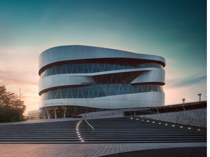 Das Mercedes-Benz Museum mit seiner modernen, geschwungenen Architektur bei Sonnenuntergang, umgeben von Treppen und einem klaren Himmel., © Mercedes-Benz Heritage