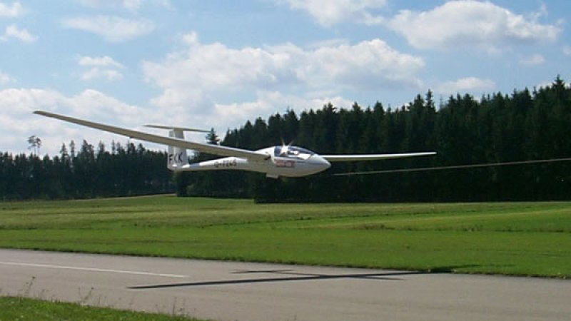 Segelflugzeug im Landeanflug auf asphaltierte Landebahn, umgeben von Wiesen und Wald. Der Himmel ist leicht bewölkt., © Aero-Club Esslingen e.V. Segelflugzeug im Landeanflug auf asphaltierte Landebahn, umgeben von Wiesen und Wald. Der Himmel ist leicht bewölkt., © Aero-Club Esslingen e.V.