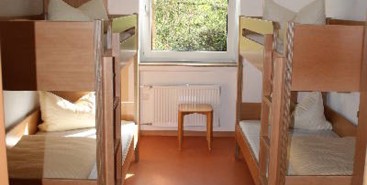 Ein heller Schlafsaal mit zwei Etagenbetten aus Holz, einem Fenster in der Mitte und einem Hocker darunter.