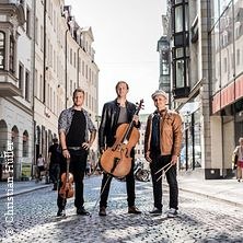 Drei Männer mit Musikinstrumenten stehen auf einer gepflasterten Straße in einer Stadt. Sie tragen lässige Kleidung und blicken in die Kamera., © links im Bild Drei Männer mit Musikinstrumenten stehen auf einer gepflasterten Straße in einer Stadt. Sie tragen lässige Kleidung und blicken in die Kamera., © links im Bild