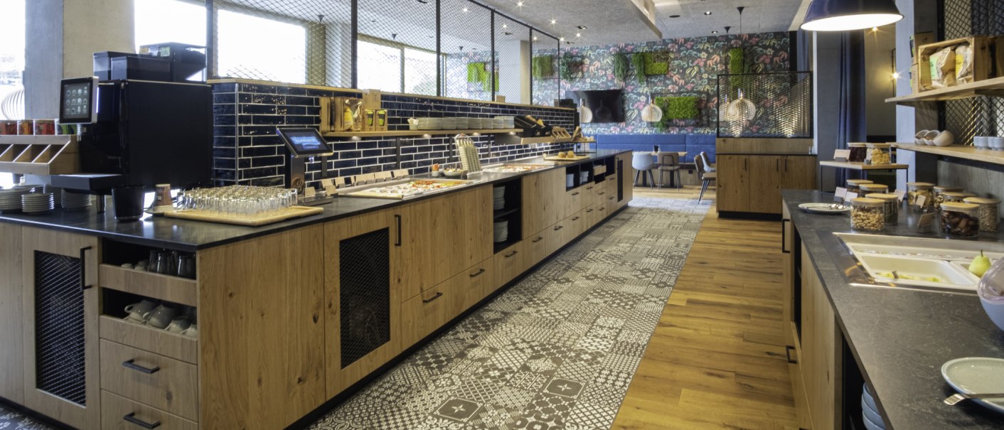 Moderner Frühstücksbereich mit Holztheken, Buffet, Kaffeemaschine und stilvollen Sitzgelegenheiten. Bunte Wanddekoration und helle Beleuchtung., © 13 6 Design GmbH (Judenhof 10, 89073 Ulm)