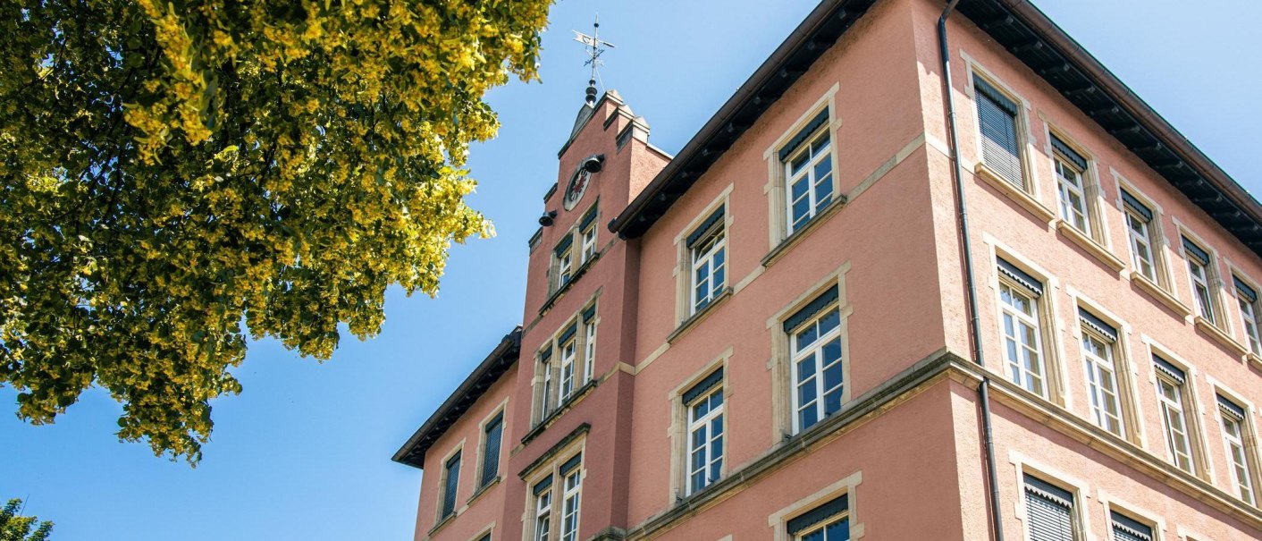 Ein rosa Gebäude mit Uhrturm und Fenstern, daneben ein Baum mit grünen Blättern unter blauem Himmel., © Stuttgart-Marketing GmbH, Sarah Schmid Ein rosa Gebäude mit Uhrturm und Fenstern, daneben ein Baum mit grünen Blättern unter blauem Himmel., © Stuttgart-Marketing GmbH, Sarah Schmid