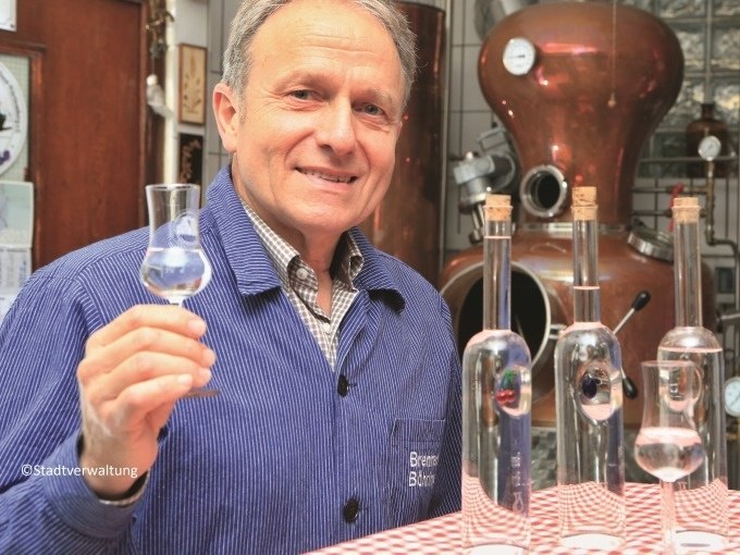 Mann in blauer Jacke hält ein Glas mit klarer Flüssigkeit vor einer Destillationsanlage. Auf dem Tisch stehen Flaschen., © Eigenbetrieb Tourismus und Citymanagement Schorndorf Mann in blauer Jacke hält ein Glas mit klarer Flüssigkeit vor einer Destillationsanlage. Auf dem Tisch stehen Flaschen., © Eigenbetrieb Tourismus und Citymanagement Schorndorf