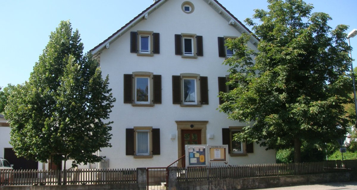 Ein weißes Haus mit braunen Fensterläden, umgeben von Bäumen und einem Holzzaun. Der Himmel ist klar und blau., © Land der 1000 Hügel - Kraichgau-Stromberg Ein weißes Haus mit braunen Fensterläden, umgeben von Bäumen und einem Holzzaun. Der Himmel ist klar und blau., © Land der 1000 Hügel - Kraichgau-Stromberg