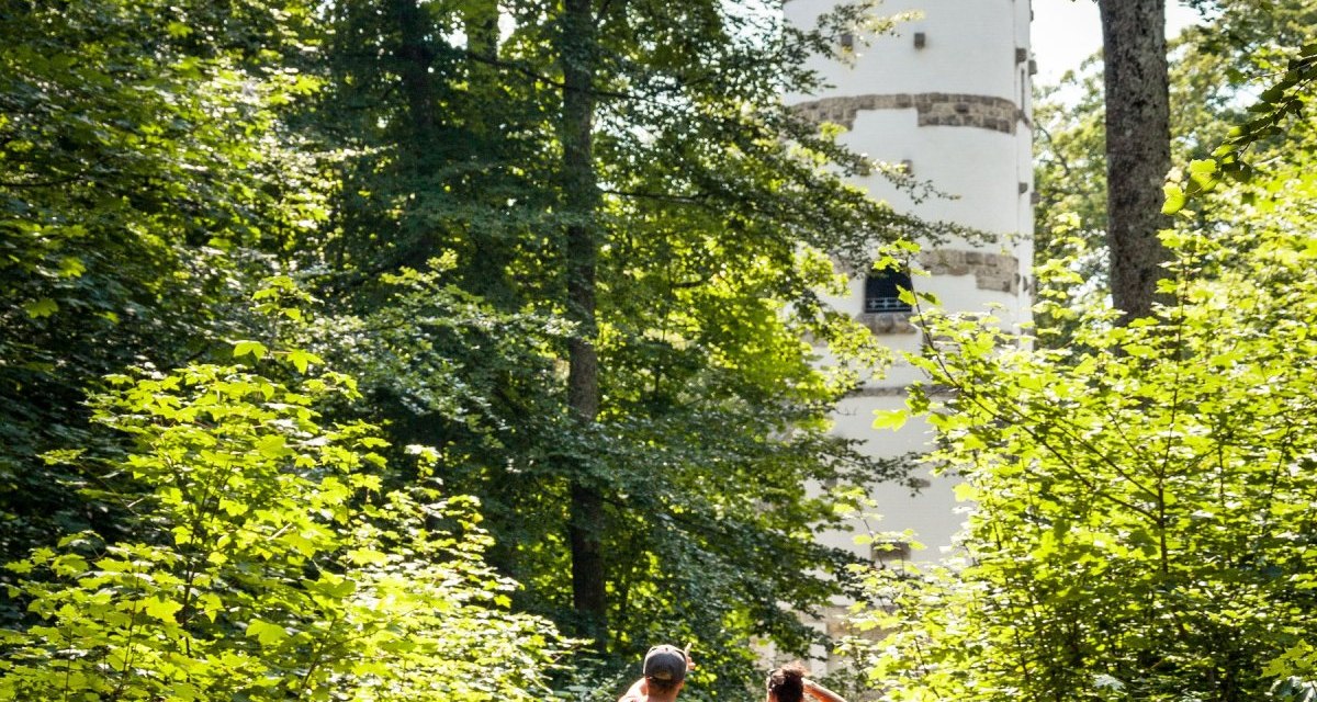 Zwei Wanderer auf einem Waldweg, umgeben von grünen Bäumen, gehen auf einen weißen Turm zu., © hochgehberge Zwei Wanderer auf einem Waldweg, umgeben von grünen Bäumen, gehen auf einen weißen Turm zu., © hochgehberge