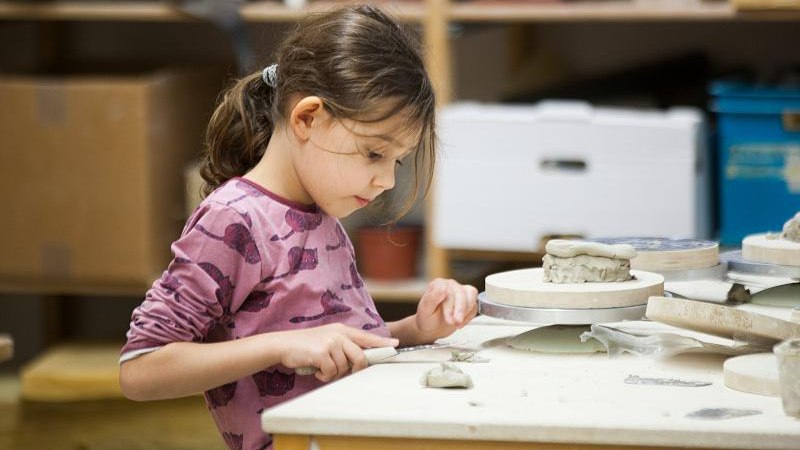 Ein Kind in einem rosa Oberteil formt konzentriert Ton auf einem Tisch in einem Workshop. Im Hintergrund sind Regale mit Materialien zu sehen., &copy; Kunstschule Labyrinth