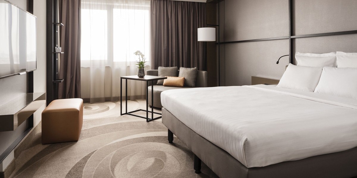 Modernes Hotelzimmer mit gro&szlig;em Bett, Sofa, Tisch und Fernseher. Helle Vorh&auml;nge und dezente Farben schaffen eine elegante Atmosph&auml;re., &copy; Stuttgart Marriott Hotel Sindelfingen