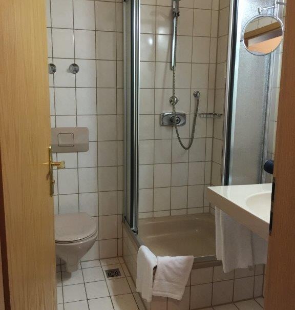 Kleines Badezimmer mit Dusche, Toilette und Waschbecken. Weiße Fliesen, Handtuch an der Duschkabine, runder Spiegel an der Wand., © TOMAS Kleines Badezimmer mit Dusche, Toilette und Waschbecken. Weiße Fliesen, Handtuch an der Duschkabine, runder Spiegel an der Wand., © TOMAS