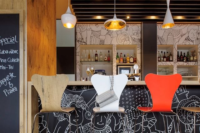 Bar mit bunten Stühlen, dekorativer Wand und Tafel mit Getränkespezialitäten. Moderne Lampen und Flaschen im Hintergrund., © TOMAS