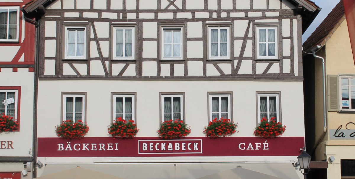 Fachwerkhaus mit Bäckerei und Café Beckabeck. Rote Blumen schmücken die Fenster. Vor dem Gebäude stehen Tische und Stühle unter Sonnenschirmen.