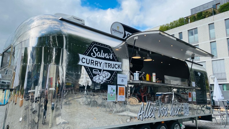 Ein glänzender Foodtruck mit der Aufschrift 'Salva's Curry Truck' steht im Freien vor einem modernen Gebäude., © Salva's Curry-Truck