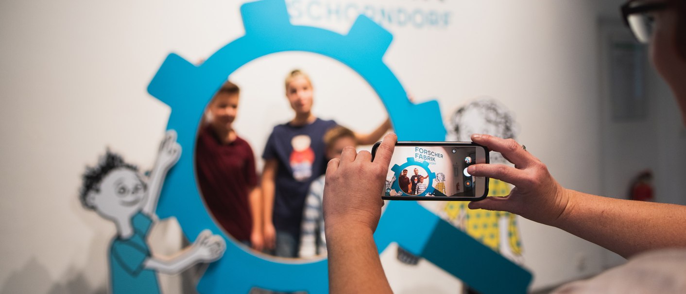Eine Person fotografiert drei Kinder durch einen blauen, zahnradförmigen Rahmen mit einem Smartphone. Im Hintergrund sind Zeichnungen zu sehen., © Danijel Grbic Eine Person fotografiert drei Kinder durch einen blauen, zahnradförmigen Rahmen mit einem Smartphone. Im Hintergrund sind Zeichnungen zu sehen., © Danijel Grbic